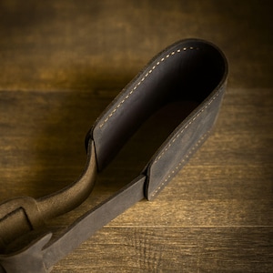 May include: A dark brown leather holder with a rounded, open-top cylindrical shape and a flat strap extending from the base. The leather features visible light-colored stitching along the edges, creating a structured and durable appearance.