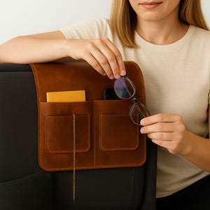 Puede incluir: Un organizador de sillón de cuero marrón con múltiples bolsillos. El organizador está diseñado para colgar sobre el brazo de una silla y contiene un cuaderno, un teléfono y un par de gafas. El organizador es de cuero marrón y tiene una cuerda dorada decorativa.