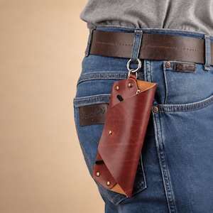May include: A reddish-brown leather glasses case with a folded envelope-style design, secured by metallic rivets. The case features a silver-toned metal ring attached to the top, allowing it to hang from a dark brown leather belt on blue jeans.