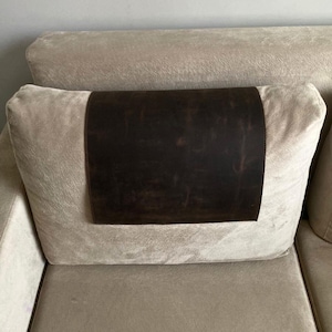 Op de afbeelding: Een rechthoekige hoofdsteunhoes van donkerbruin leer met een gladde, matte afwerking en zichtbare natuurlijke nerf, centraal geplaatst over een lichtbeige, zacht aanvoelend stoffen kussen met een pluizig, licht geborsteld uiterlijk.