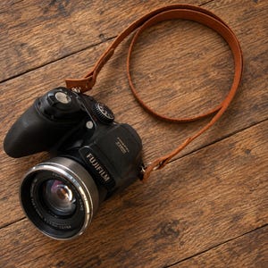 Könnte beinhalten: Eine schwarze Fujifilm FinePix S8600 Digitalkamera mit silbernem Objektivring und einem hellbraunen Leder-Trageriemen. Die Kamera verfügt über einen ausgeprägten Griff, ein Modus-Wahlrad auf der Oberseite und ein Objektiv mit sichtbarer Beschriftung.
