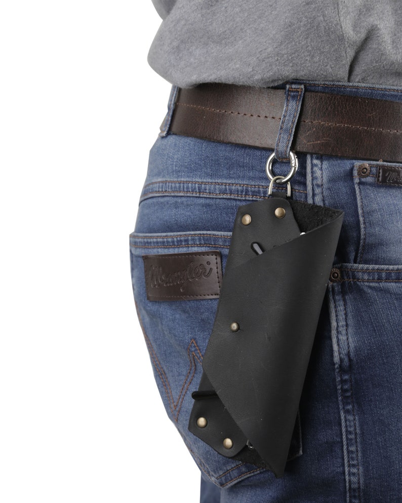 May include: A black leather phone case with a metal clip attached to a brown leather belt. The case is hanging from the belt loop of a pair of blue jeans.