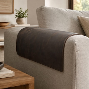 Può includere: Un copribracciolo in pelle marrone scuro drappeggiato sul bracciolo di un divano imbottito color beige chiaro. La copertura rettangolare presenta una finitura liscia e opaca con cuciture visibili lungo i bordi, progettata per poggiare piatta sul bracciolo.