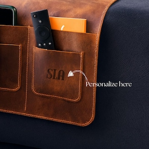May include: A brown leather couch organizer with multiple pockets. A remote control, a phone, and a small orange book are visible in the pockets. The organizer has the initials "SLA" and the text "Personalize here" on the front.