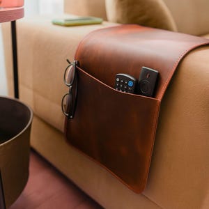 Puede incluir: Un organizador de reposabrazos de sofá de cuero marrón con un bolsillo. El bolsillo contiene dos mandos a distancia y un par de gafas. El organizador está colocado sobre el brazo de un sofá beige.