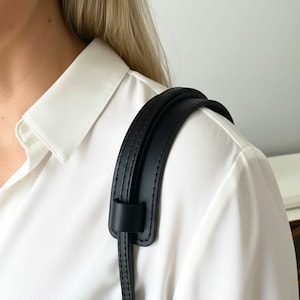 May include: A black leather shoulder strap with a padded design. The strap is attached to a black leather loop. The strap is worn over a white collared shirt.