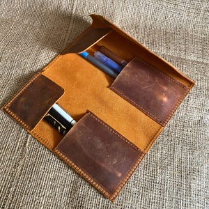 May include: A brown leather pencil case with three slots for pens and pencils. The case is open and the slots are visible. The case is made of distressed leather and has a brown suede lining.