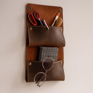May include: A brown leather wall organiser with two pockets featuring metal rivets. The top pocket holds scissors, pens, and a brush. The bottom pocket contains a small black notebook with white geometric patterns and a pair of thin-rimmed glasses.