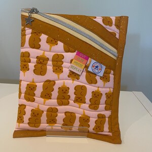 May include: A quilted fabric pouch with a zipper closure and a star-shaped charm. The pouch features a repeating pattern of brown bear-shaped designs on a pink background. The edges are trimmed with a brown fabric.