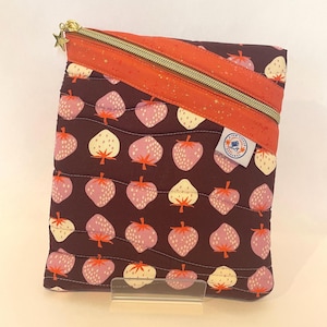 May include: A quilted pouch with a repeating strawberry pattern in pink and cream on a dark burgundy background. The pouch has an orange zipper and a gold star zipper pull. A small label is visible on the front.