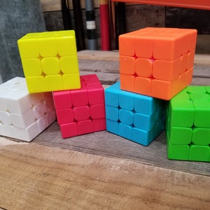 May include: Six colorful 3x3 cube puzzles, in white, yellow, orange, red, blue, and green, arranged in a pyramid shape on a wooden surface.