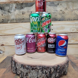 May include: A collection of seven 12-ounce aluminum soda cans, including Dr Pepper, Diet Dr Pepper, Cherry Coke, Mountain Dew Code Red, Mountain Dew, Sprite, and Pepsi. The cans are arranged in a pyramid shape on a wooden tree stump.