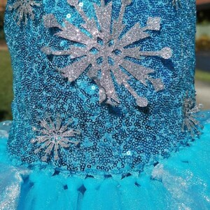 May include: A bright blue dress bodice covered in shimmering sequins, featuring a large central silver glitter snowflake appliqu&eacute; and smaller silver glitter snowflake accents scattered across the front surface above a layered tulle skirt.
