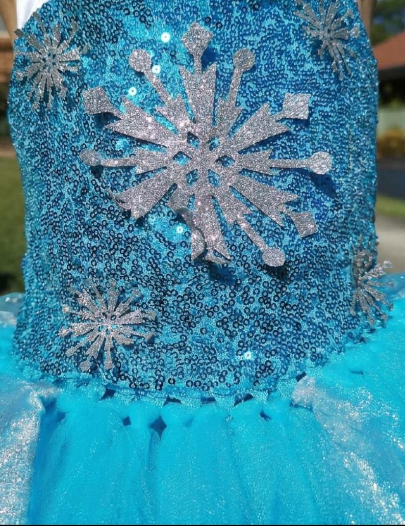 May include: A bright blue dress bodice covered in shimmering sequins, featuring a large central silver glitter snowflake appliqu&eacute; and smaller silver glitter snowflake accents scattered across the front surface above a layered tulle skirt.