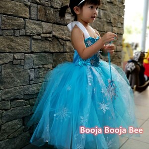 May include: A bright blue sleeveless dress with a sequined bodice and a layered tulle skirt featuring silver snowflake patterns. The dress includes white halter straps and is paired with a silver tiara with blue accents. Text reads Booja Booja Bee.