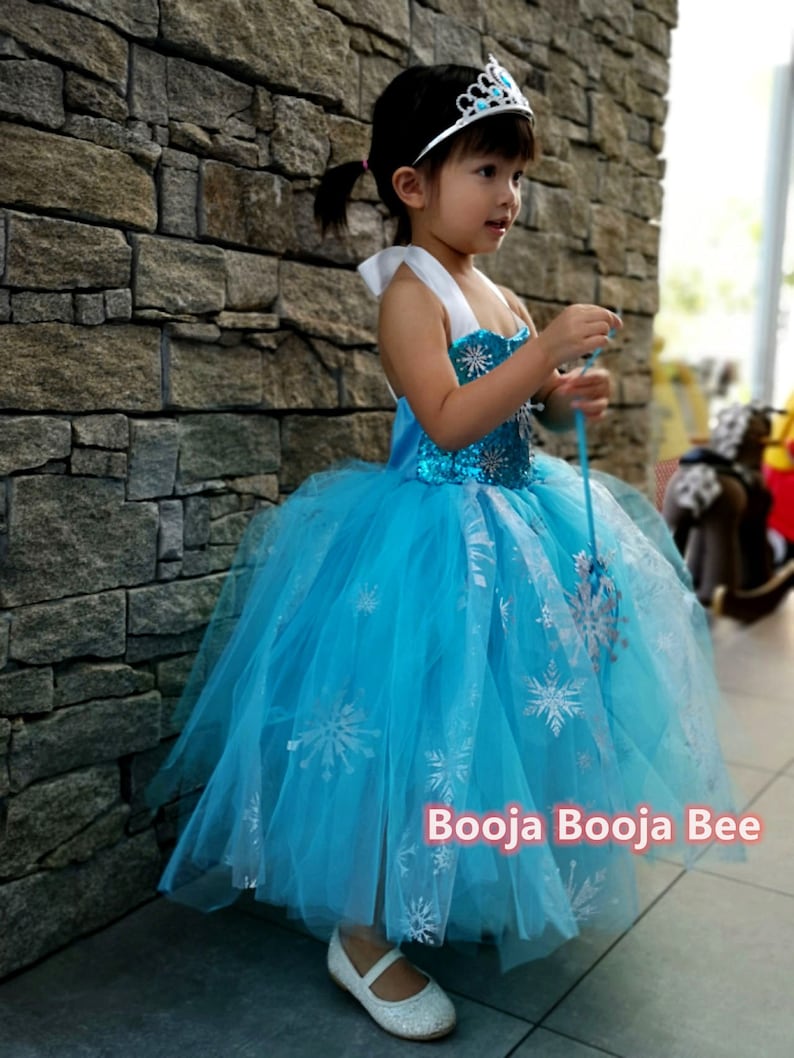 May include: A bright blue sleeveless dress with a sequined bodice and a layered tulle skirt featuring silver snowflake patterns. The dress includes white halter straps and is paired with a silver tiara with blue accents. Text reads Booja Booja Bee.