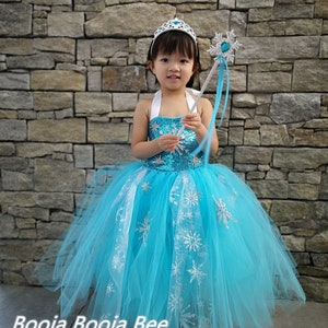 May include: A bright blue sequined halter dress with a full tulle skirt featuring silver snowflake patterns. The outfit includes a silver tiara with a blue gem and a matching silver snowflake wand with blue ribbons. The text Booja Booja Bee is visible.