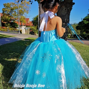 May include: A bright blue halter-style dress featuring a full, layered tulle skirt adorned with silver snowflake patterns. The bodice includes a white satin ribbon tie at the neck. The text Booja Booja Bee is visible at the bottom of the image.