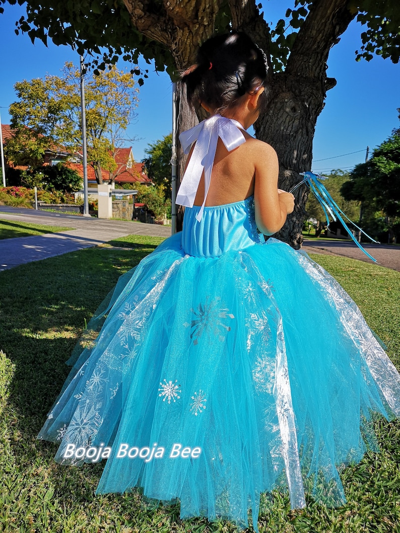 May include: A bright blue halter-style dress featuring a full, layered tulle skirt adorned with silver snowflake patterns. The bodice includes a white satin ribbon tie at the neck. The text Booja Booja Bee is visible at the bottom of the image.