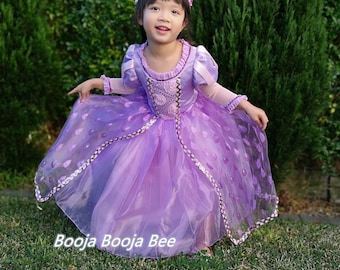 May include: A young child wearing a lavender princess dress with a floral crown. The dress features a bodice with floral accents, long sleeves, and a layered skirt with a decorative trim. The text "Booja Booja Bee" is visible on the dress.