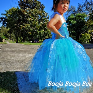 May include: A bright blue sleeveless dress featuring a sequined bodice and a layered tulle skirt adorned with silver snowflake patterns. The dress includes white shoulder straps and is paired with a silver-toned tiara with a central blue gem.