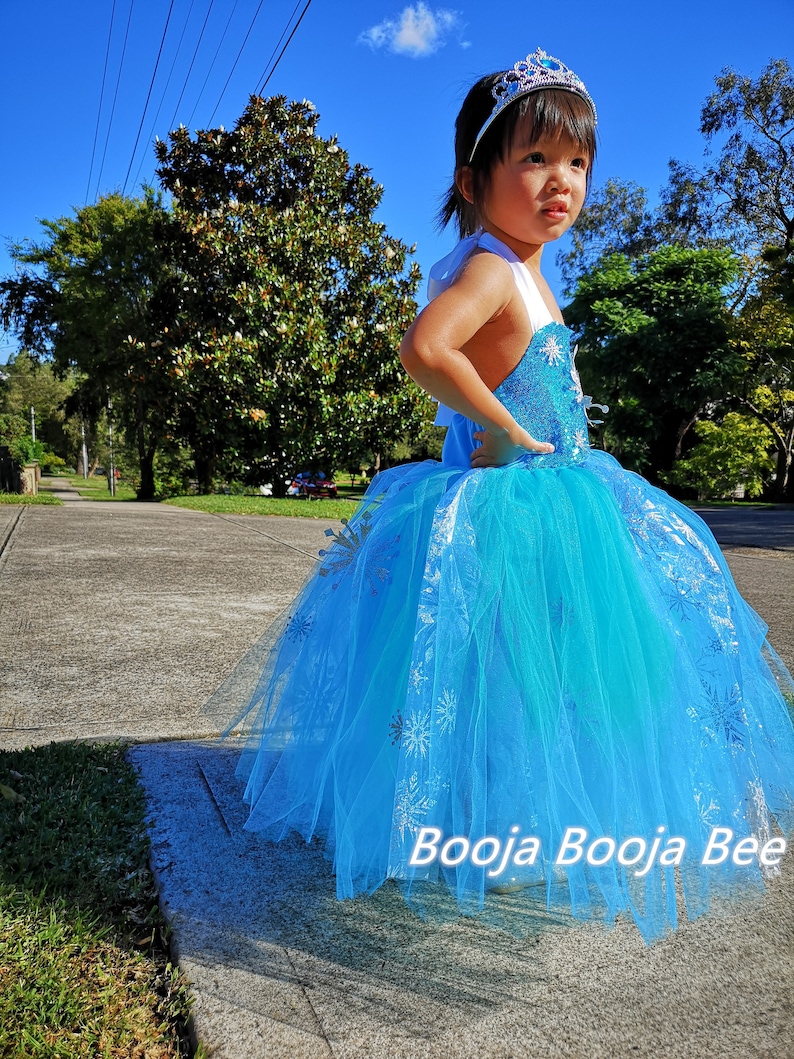 May include: A bright blue sleeveless dress featuring a sequined bodice and a layered tulle skirt adorned with silver snowflake patterns. The dress includes white shoulder straps and is paired with a silver-toned tiara with a central blue gem.