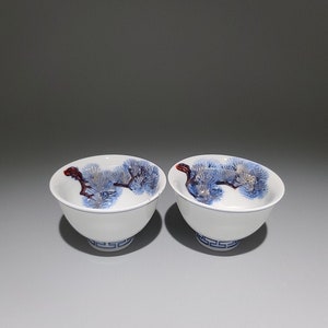May include: Two white ceramic teacups with blue and red pine tree designs. The cups have a blue and white geometric pattern around the base.