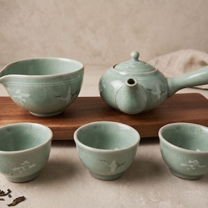 May include: A celadon green tea set featuring a side-handled teapot, a pouring pitcher, and three small tea cups. Each piece displays a delicate crackle glaze finish and white crane and pine tree motifs painted on the exterior surfaces.