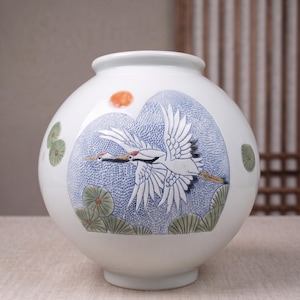 May include: A white ceramic vase with a spherical body featuring a hand-painted scene of a crane in flight. The design includes a blue stippled background, a small orange sun, and green lily pads. The vase has a smooth, glossy finish and a wide opening.