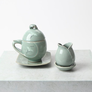 May include: A celadon-colored ceramic tea set featuring a lidded cup with a handle, a small tulip-shaped vase, and a square saucer. The cup and lid are adorned with crane illustrations. The set is displayed on a textured surface.