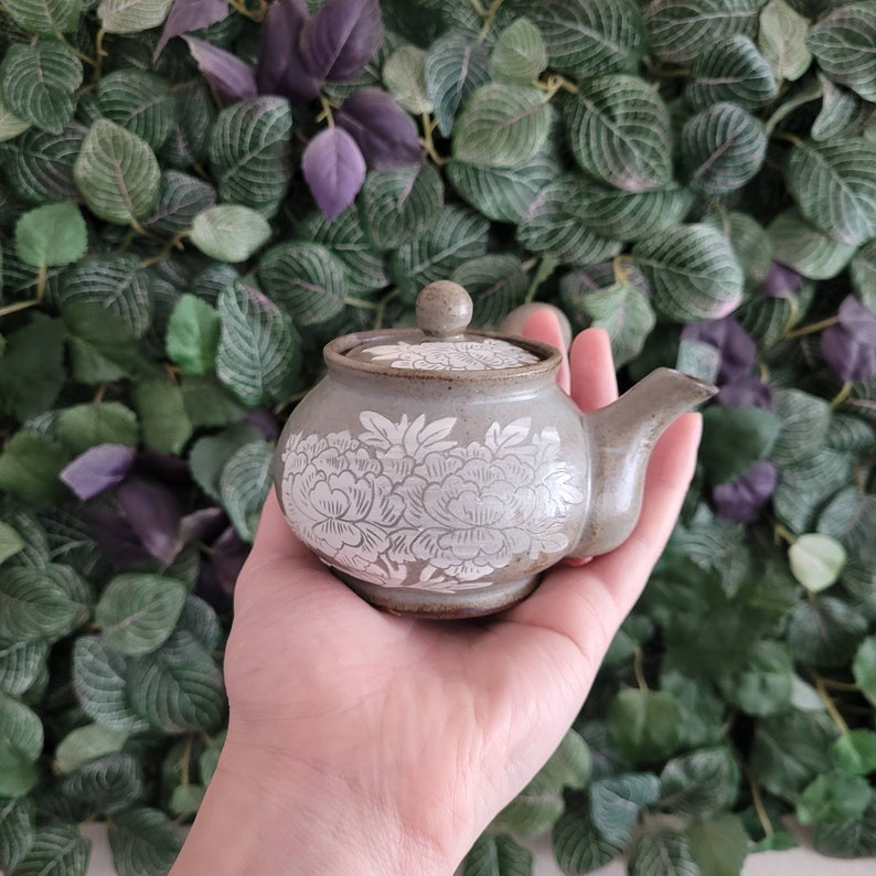 May include: A small, gray ceramic teapot with a white floral design. The teapot has a lid and a spout. It is sitting on a hand.