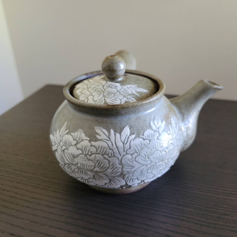 May include: A gray ceramic teapot with a white floral design. The teapot has a lid and a spout.