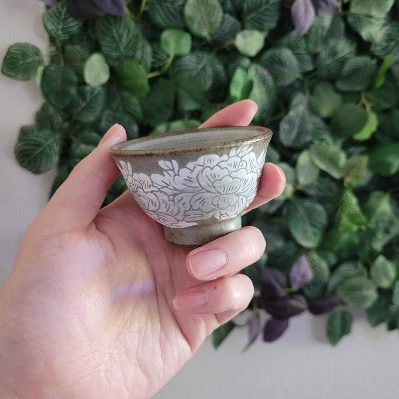 May include: A small, gray ceramic teacup with a white floral design. The cup is held in a hand.