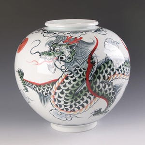 May include: White ceramic vase featuring a hand-painted green dragon design. The dragon is depicted with red accents and swirling clouds.