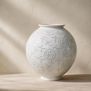 May include: A spherical white ceramic vase featuring a repeating embossed pattern of cranes in flight and circular medallions. The surface has a smooth, glossy finish with a narrow, defined rim at the top and a small, circular base at the bottom.