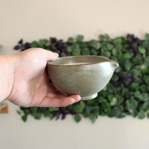 May include: A small, light gray ceramic bowl with a white liquid inside. The bowl has a slightly textured surface and a small spout on one side.