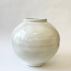 以下が含まれることがあります： 丸みを帯びたオフホワイトのセラミック花瓶。幅広の球状のボディと細い首が特徴です。光沢のある仕上がりで、小さな円形のベースに置かれています。シンプルでエレガントなデザインで、インテリアに最適です。