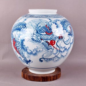 May include: A white ceramic vase with a blue and red dragon design. The vase is round and has a wide mouth. It is sitting on a wooden base.