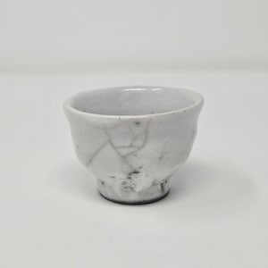 以下が含まれることがあります： ひび割れた釉薬を施した、小さな白い陶器のカップ。カップは丸みを帯びた底とわずかに広がった縁を持っています。