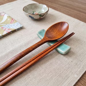 May include: A polished wooden spoon and a pair of matching wooden chopsticks resting on a light blue ceramic chopstick holder with a crackled glaze finish. A small, shallow ceramic bowl with a floral pattern sits in the background.