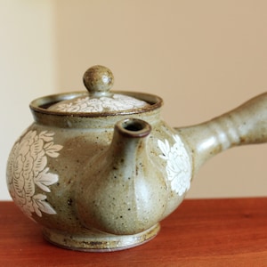 May include: A small, brown ceramic teapot with a white floral design. The teapot has a spout and a handle.