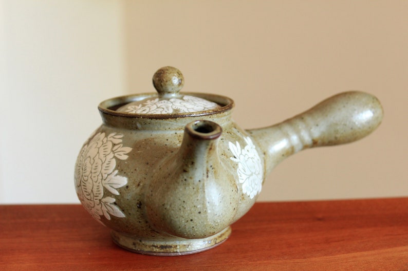 May include: A small, brown ceramic teapot with a white floral design. The teapot has a spout and a handle.