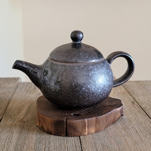May include: A black speckled ceramic teapot with a lid and a wooden base. The teapot has a rounded body and a spout on the side.