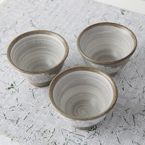 May include: Three small, white ceramic teacups with a brown rim and a swirl pattern. The cups have a floral design on the bottom.