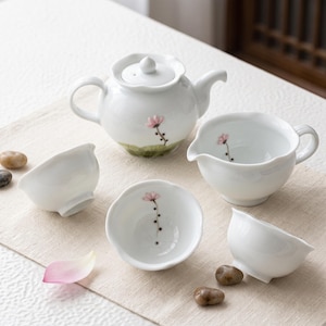 May include: A white ceramic tea set consisting of a teapot with a lid, a pitcher with a spout, and three small tea bowls. Each piece features a delicate hand-painted pink flower on a thin brown stem, with a scalloped rim design on the teapot lid.