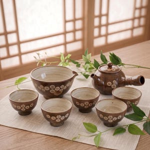 May include: A ceramic tea set featuring a brown teapot with a side handle and lid, a large mixing bowl, and five small tea cups. Each piece is decorated with a white daisy pattern and has a light-colored interior with visible horizontal lines.