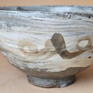 May include: A ceramic bowl with a textured, uneven surface featuring a marbled blend of grey, white, and beige tones. The exterior displays a hand-painted, swirling brown brushstroke design that wraps around the side of the rounded vessel.