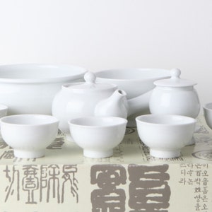 Op de afbeelding: Een witte porseleinen theeset met een theepot, een suikerpot met deksel en zes kopjes. De set is gerangschikt op een gepatroneerd oppervlak.