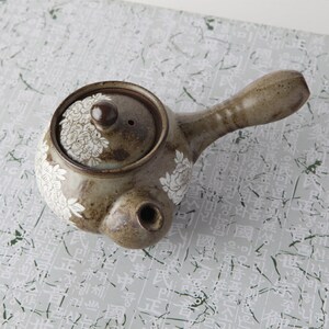 May include: A brown ceramic teapot with a white floral design. The teapot has a small spout and a handle. The lid has a small knob on top.