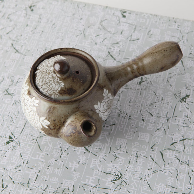 May include: A brown ceramic teapot with a white floral design. The teapot has a small spout and a handle. The lid has a small knob on top.
